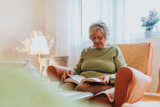 A mature woman enjoying a book in a comfortable armchair, surrounded by a warm and inviting atmosphere with soft lighting and homely decor. - Powered by Adobe