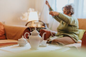 Senior woman sitting on couch in a warmly lit living room, knitting while a porcelain tea set sits elegantly in the foreground, symbolizing tranquility and warm moments.