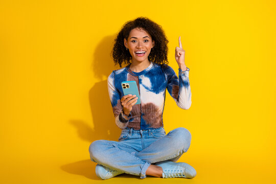 Young stylish woman with curly hair holding a smartphone against a vibrant yellow background, expressing enthusiasm.