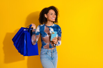 Young stylish woman holding a smartphone and shopping bags against a vibrant yellow background,...