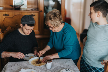 A senior woman helps prepare a pancake while two young men sit at the dining table, fostering...