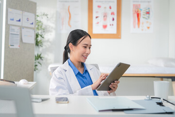 Portrait of a confident Asian female doctor smiling while working at her desk in a clinic, typing on her laptop and reviewing medical reports. Modern healthcare professional indoors with document file