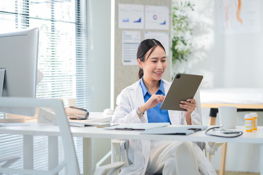 Portrait of a confident Asian female doctor smiling while working at her desk in a clinic, typing on her laptop and reviewing medical reports. Modern healthcare professional indoors with document file