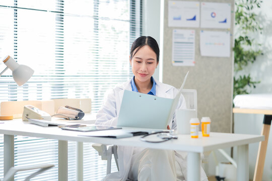 Portrait of a confident Asian female doctor smiling while working at her desk in a clinic, typing on her laptop and reviewing medical reports. Modern healthcare professional indoors with document file