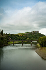 A tranquil river flows beneath a modest concrete bridge, framed by lush green hills and dramatic...