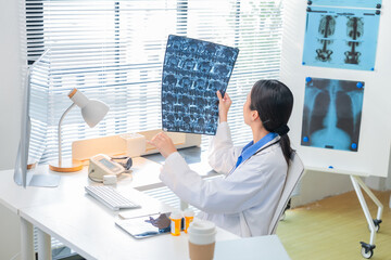 Asian female doctor or nurse in a white lab coat analyzing an X-ray scan in a medical clinic. She examines patient radiographs carefully, studying brain or lung images on film or screen, representing 