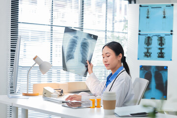 Asian female doctor or nurse in a white lab coat analyzing an X-ray scan in a medical clinic. She examines patient radiographs carefully, studying brain or lung images on film or screen, representing 