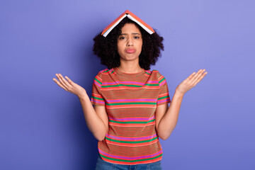 Worried young woman with curly hair holding up a book on her head and demonstrating a confused...