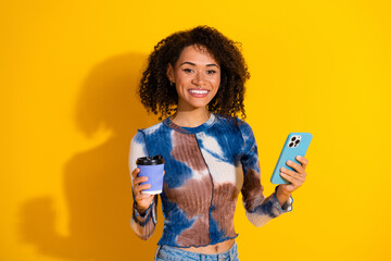 Stylish young woman with curly hair holding a phone and coffee cup, smiling brightly against a...