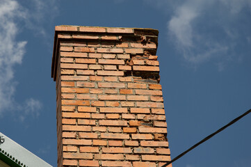 Chimney displays significant wear and damage with cracks against a backdrop of a clear blue sky, indicating possible structural concerns