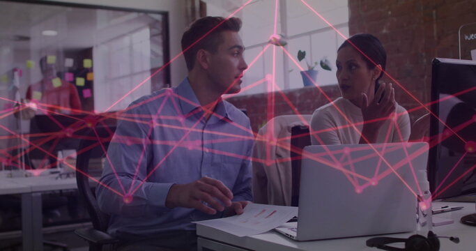 Pointing colleagues discussing data flow at office desk with sticky notes, showing laptop printouts