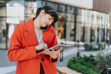 A businesswoman in a striking red coat, multitasking with a tablet and smart phone outdoors in a...