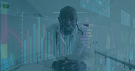 Leaning mature doctor wearing white coat in clinic corridor, with stethoscope and digital charts