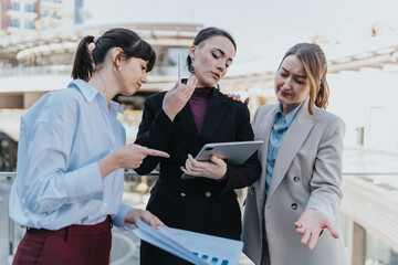 Three women, dressed professionally, engage in an outdoor business discussion involving documents...