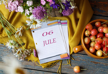 Calendar for July 6: flip calendar with numbers 06, month name July in English on a yellow napkin with wild flowers and ripe cherries