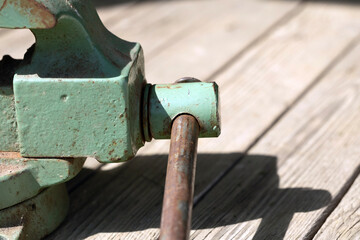 A close-up of a green metal clamp on a wooden surface. The clamp features a rusted screw mechanism...
