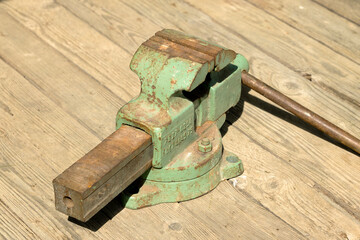 A green metal vise is mounted on a wooden surface. The vise has a sturdy design with a wooden...