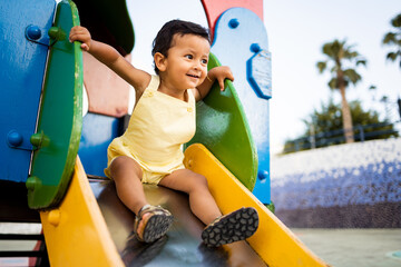 A young Latino boy between 1 and 2 years old in casual clothes is playing in a park at sunset.The child is happily going down a slide. Concept of Latino babies.
