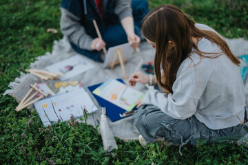 Two teenagers are outdoors in a green park, painting and sketching together. They are sitting on a...