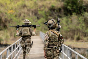 Two armed special forces soldiers in full tactical gear patrolling a narrow bridge during field...