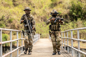 Two armed special forces soldiers in full tactical gear patrolling a narrow bridge during field...