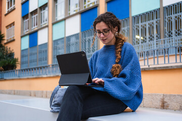 Focused female student working on tablet in front of colorful modern building, enjoying outdoor study session