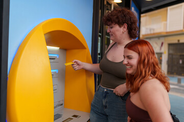 Two friends withdrawing money from a yellow ATM, one inserting the card and the other laughing