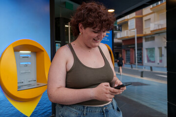 Smiling young transgender woman using smartphone in front of bank office, online banking and...
