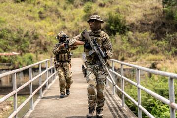 Two armed special forces soldiers in full tactical gear patrolling a narrow bridge during field...