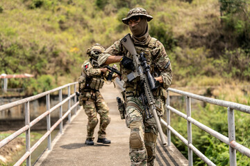 Two armed special forces soldiers in full tactical gear patrolling a narrow bridge during field...