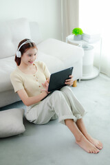 An asian woman is sitting against the sofa, using a tablet to watch entertainment at home