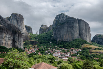 Spectacular view of the famous Meteora rock formations rising above a traditional Greek village. Dramatic cliffs covered with greenery under a cloudy sky, showcasing the unique landscape.