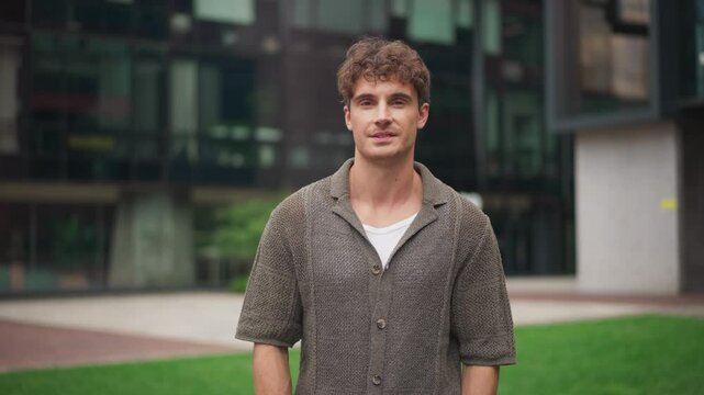 Portrait of young Caucasian man standing outdoors in front of modern office building. Handsome male wearing casual knitted shirt and white undershirt posing near green lawn in business district.