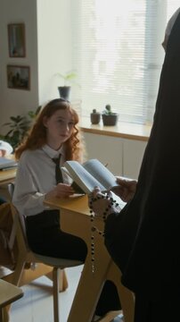 Vertical of Caucasian teen girl in white uniform sitting at single desk listening carefully to unrecognizable nun in veil reading Bible out loud during catechism class in Catholic school