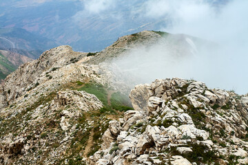 An imposing, heavily weathered rock cliff with debris. Light tones indicate the rock type. A mountainous landscape of Tashkent Region, Uzbekistan, partly hidden in mist