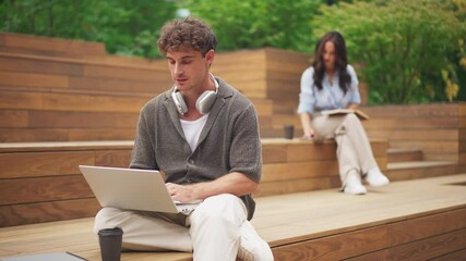 Cheerful man with headphones using laptop and chatting on wooden steps, woman in background reading. Productive atmosphere outdoors. Male student texting and smiling while break at work or study. - Powered by Adobe