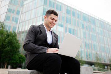 Professional young man working on laptop outdoors in a modern urban setting surrounded by greenery...