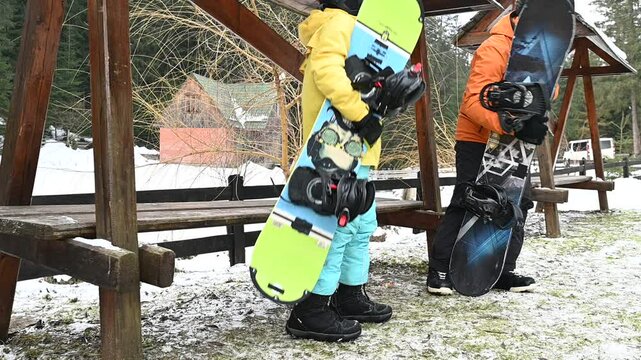Two snowboarders pause on a snowy day, their boards leaning against a rustic wooden bench. A peaceful winter scene.