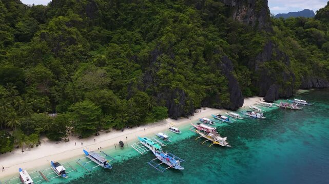 Long clip from close up to wide Aereal view of Colorful Bangka Boats Docked on the Crystal-Clear Waters and Coral Reefs of Entalula Island with a beautiful beach with people in El Nido, Palawan, Phili
