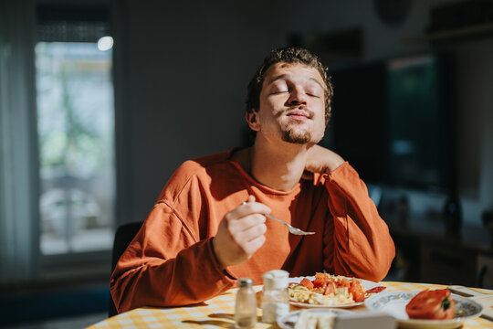 A young man wearing an orange sweater delighting in a meal served on a checkered table. Warm light emphasizes his relaxed ambiance in a cozy and modern indoor setting.