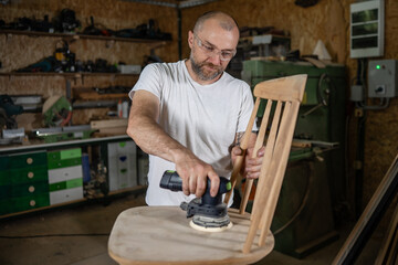 Carpenter restoring wooden chair with orbital sander in workshop