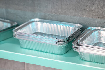 Multiple sizes of disposable aluminum food containers displayed neatly on a turquoise shelf with a gray textured wall in the background