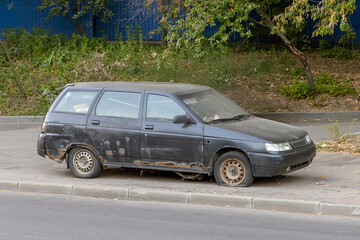 Old damaged black station wagon with rust and flat tires parked on urban pavement, surrounded by trees and fallen leaves in autumn