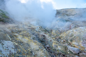 Hot steam rising from colorful geothermal landscape, rocky terrain with minerals visible, clear blue sky in background, showcasing natural geothermal activity