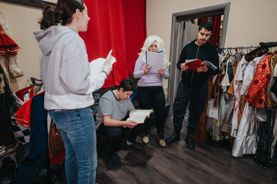 A group of young thespians standing in a dressing room filled with costumes, deeply engaged in script rehearsal and preparing for an upcoming performance.