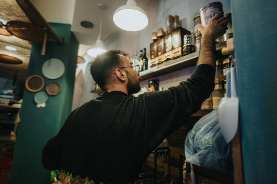 A bearded man selects a product from a wooden shelf in a warmly lit shop.