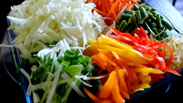 Colorful mix of freshly sliced vegetables including cabbage, carrots, bell peppers, spring onions, and green beans arranged in a glass container, ideal for salads or stir-fry.