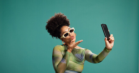 Selfie, blowing kiss and woman in studio, picture and social media on green background. Person, space and model with emoji, photography and content creation with confidence, sunglasses and influencer