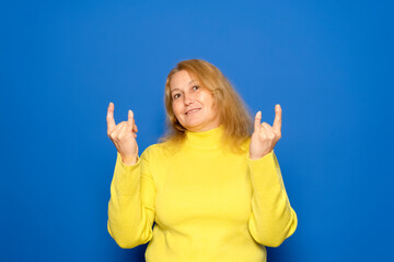 Delighted young ethnic female millennial with long dark hair in warm sweater and sunglasses, smiling brightly and showing rock gesture while listening to favorite song against yellow background