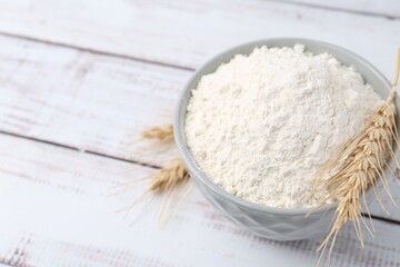 Wheat flour in bowl and spikes on light wooden table, above view. Space for text